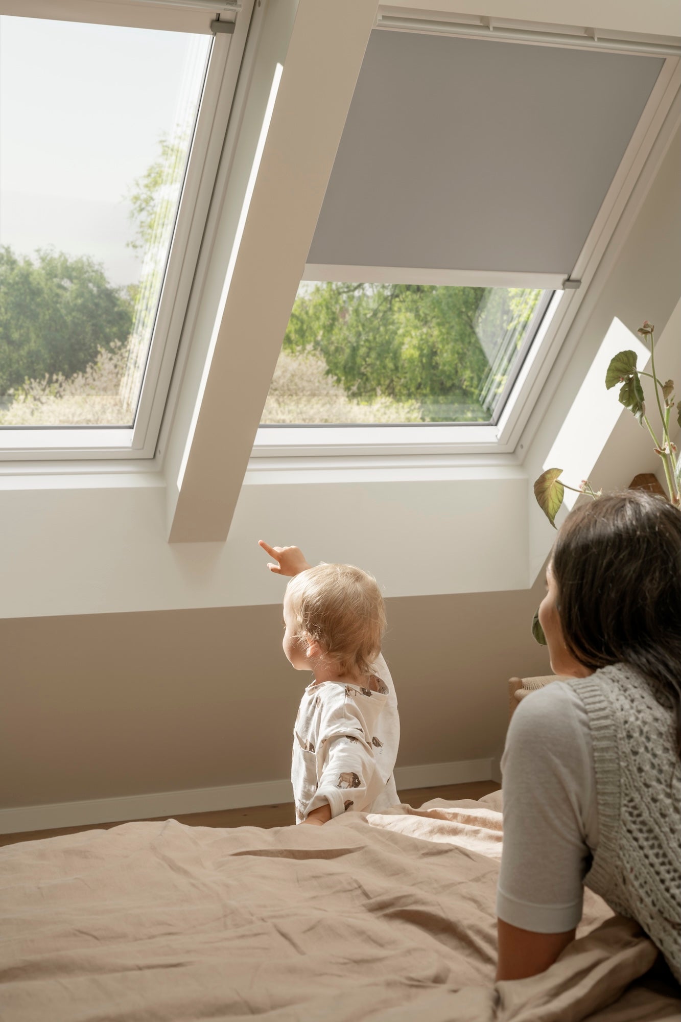 Woman and child sitting on a bed looking out a large window with a view of trees.