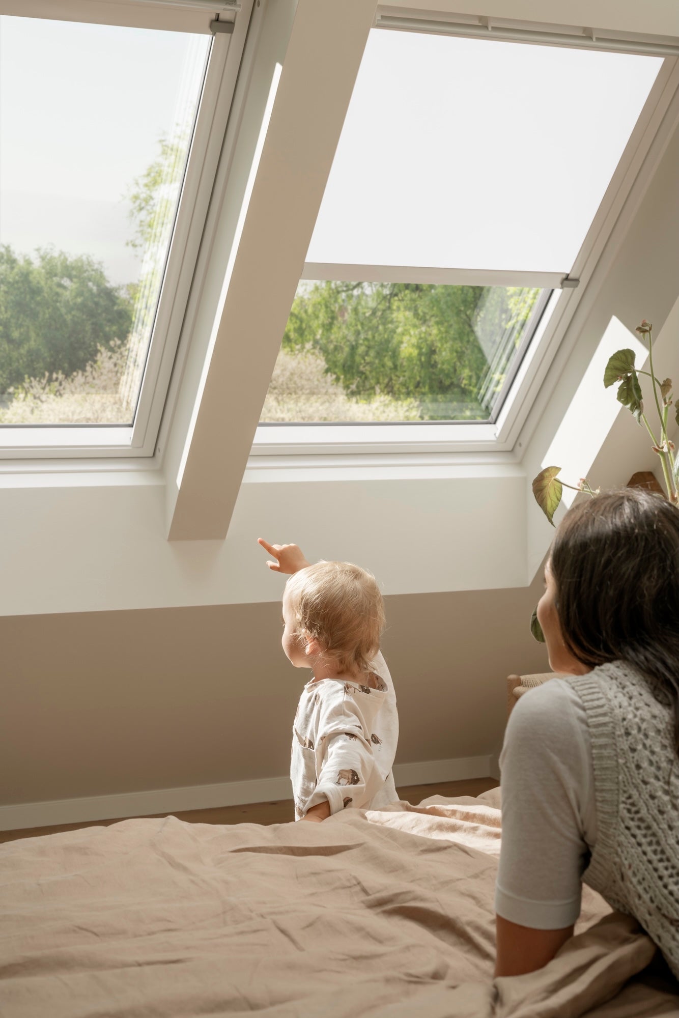 Woman and child sitting on a bed looking out a large window with blinds.