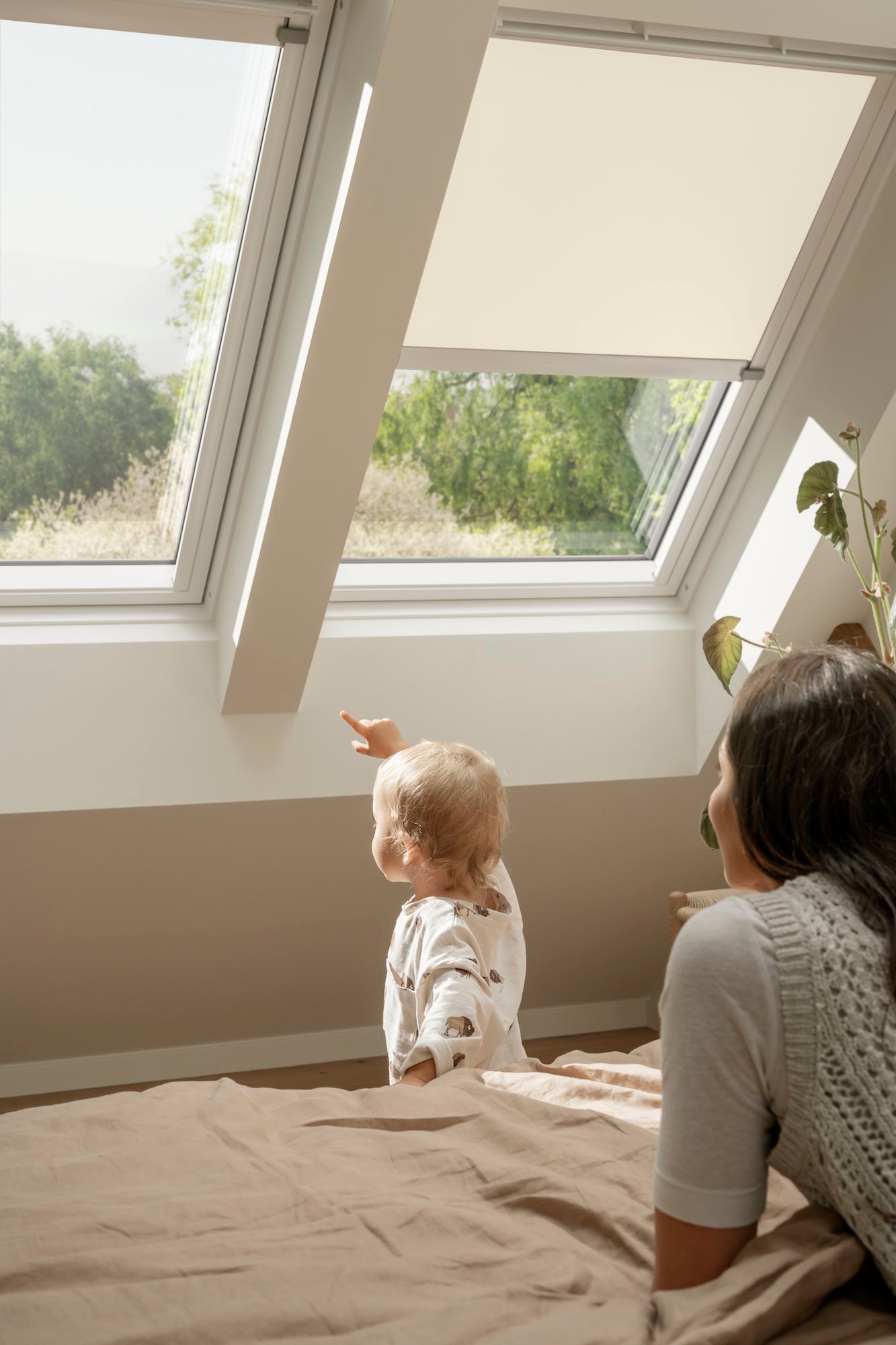Woman and child sitting on a bed looking out a large window with a view of trees.