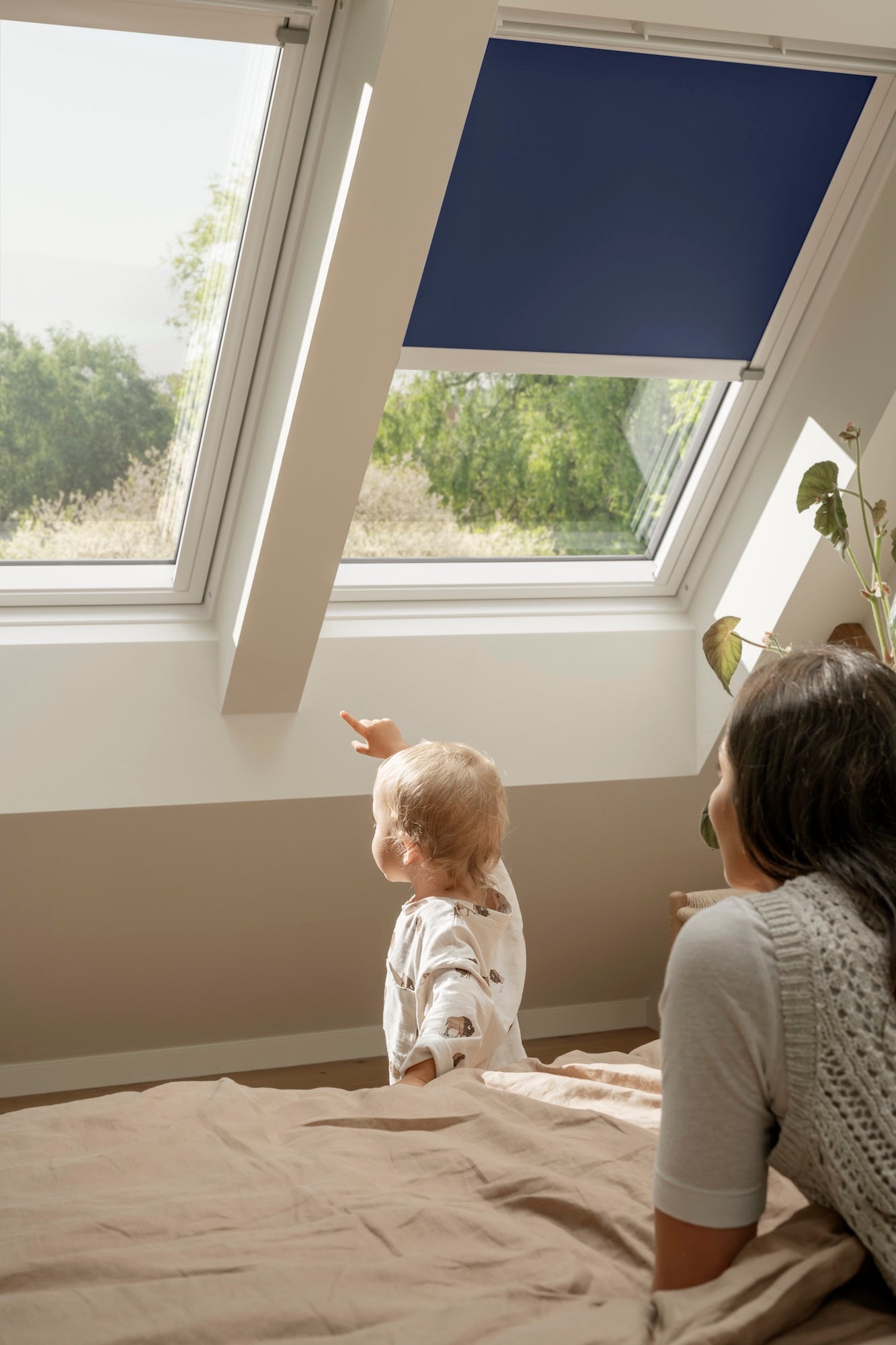 Woman and child sitting on a bed looking out a window with a blue blind.